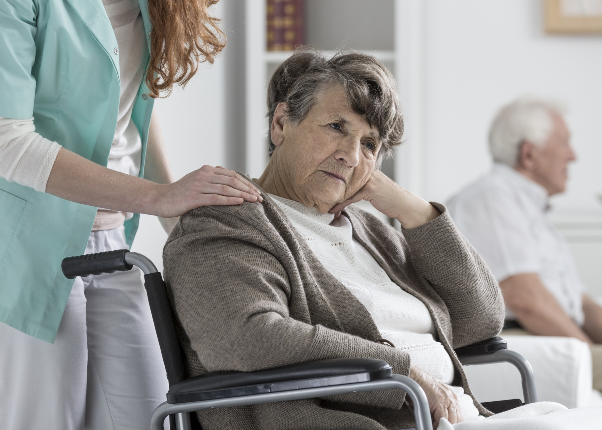 A depressed elderly woman is seated in a wheelchair with her caregiver