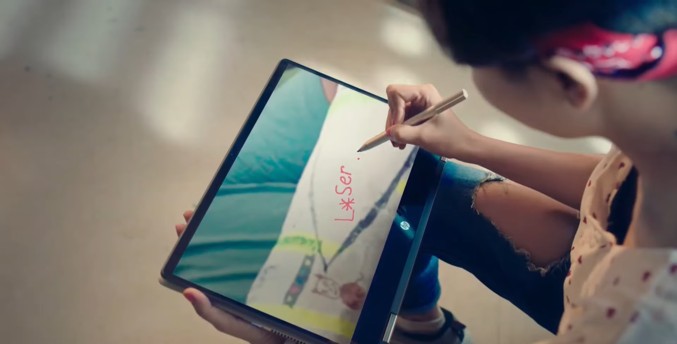 An adult woman scribbling the word ‘L*ser’ on an HP Pavilion x360 touchscreen laptop using a stylus