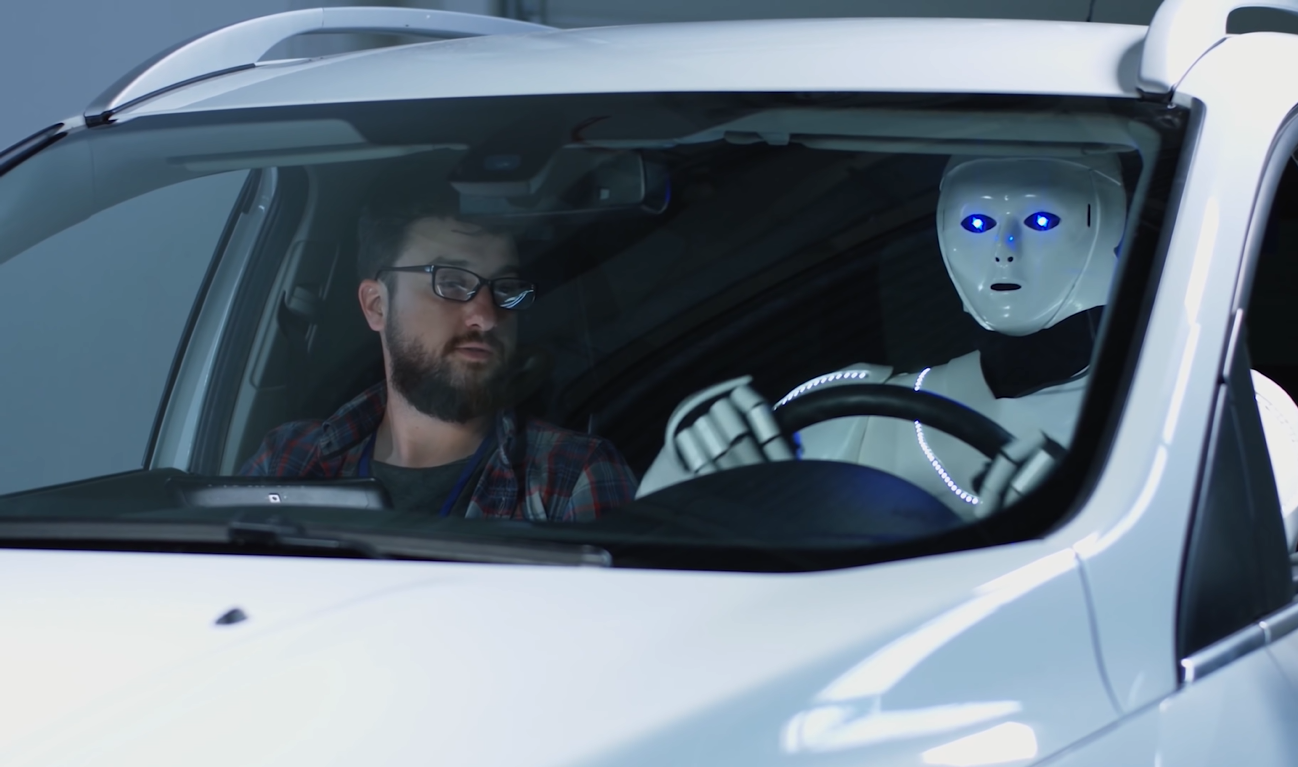 Bipedal humanoid robot ATLAS inside a car behind the wheel with an adult male on the passenger seat