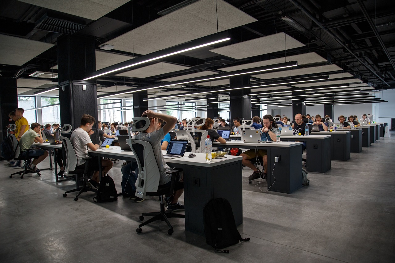 People mostly male and in T-shirt and shorts working on a laptop in a spacious and high-ceilinged office